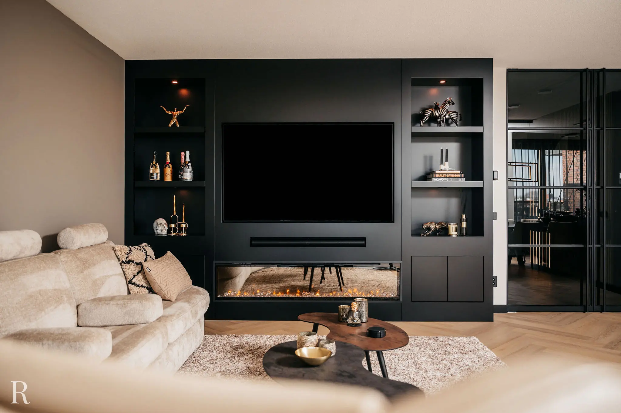 Moderne woonkamer met een zwart wandmeubel met ingebouwde tv, planken met decor en flessen en een horizontale elektrische open haard eronder. Een beige bank en salontafel staan op de voorgrond.