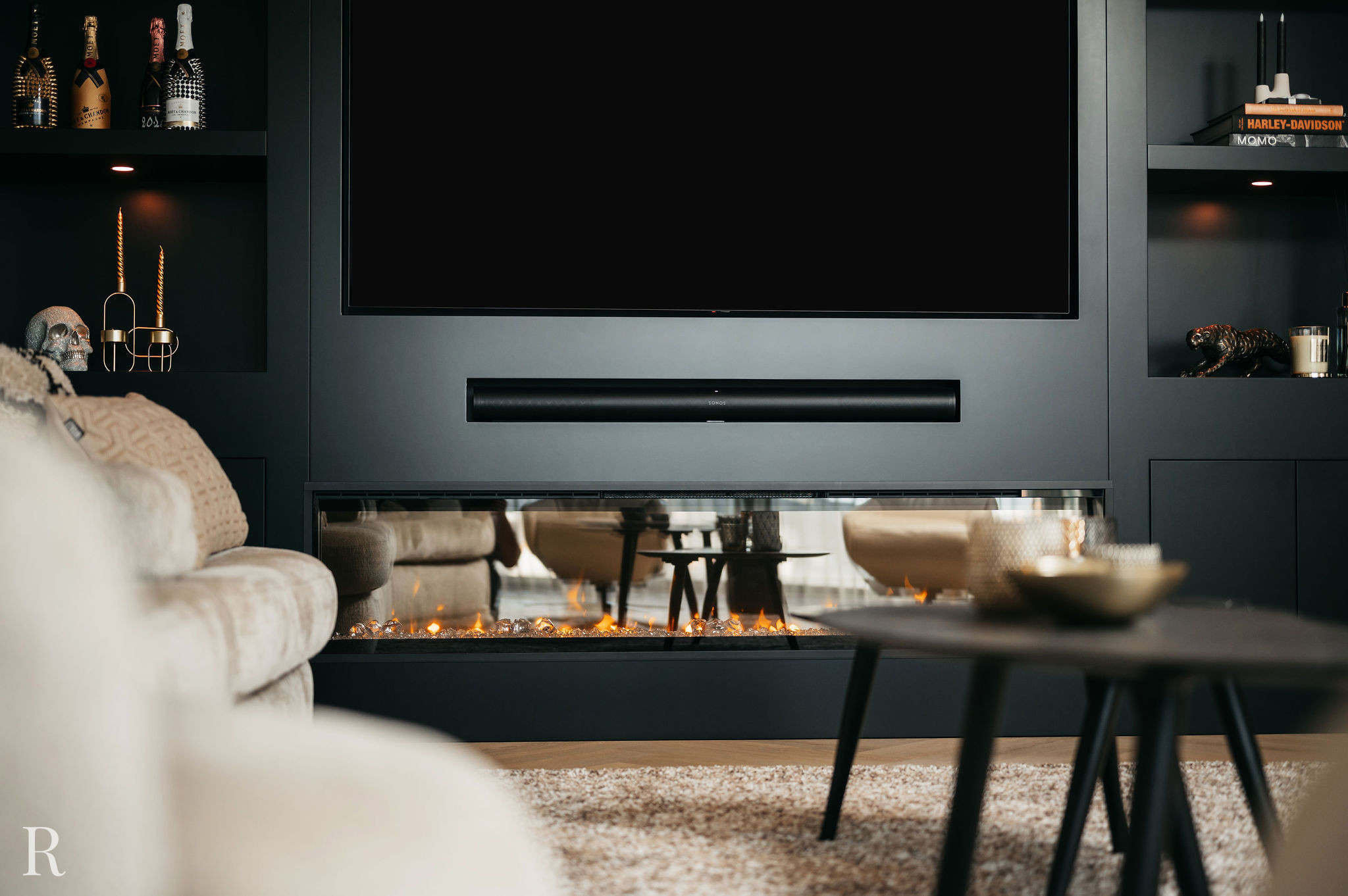 Moderne woonkamer met een zwart wandmeubel, grote tv, decoratieve rekken en een lineaire elektrische open haard eronder. Een pluche beige bank, ronde salontafel en tapijt met structuur voegen warmte toe aan de moderne ruimte.