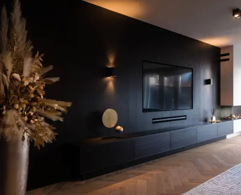 Moderne woonkamer met een zwarte accentmuur met een gemonteerde tv, minimale planken, decoratieve items en warme sfeerverlichting. Een vaas met gedroogde planten staat links op de houten vloer.