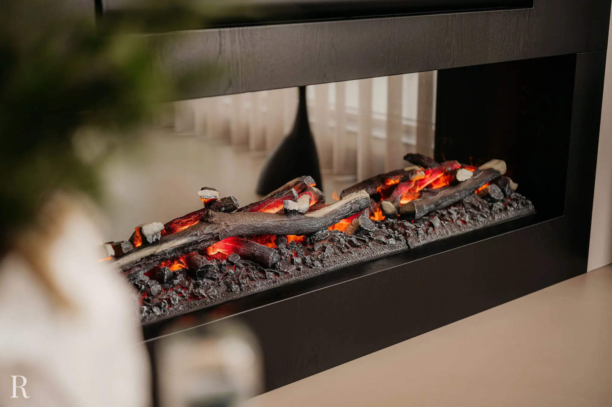 Een close-up van een moderne elektrische open haard met realistisch ogende houtblokken en gloeiende sintels, omlijst door een strakke zwarte rand. Het warme, flikkerende licht creëert een gezellige sfeer in de kamer.