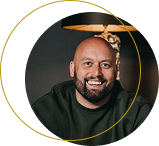 Glimlachende kale man met een baard die een donker shirt draagt, zittend binnen met een lamp op de achtergrond.