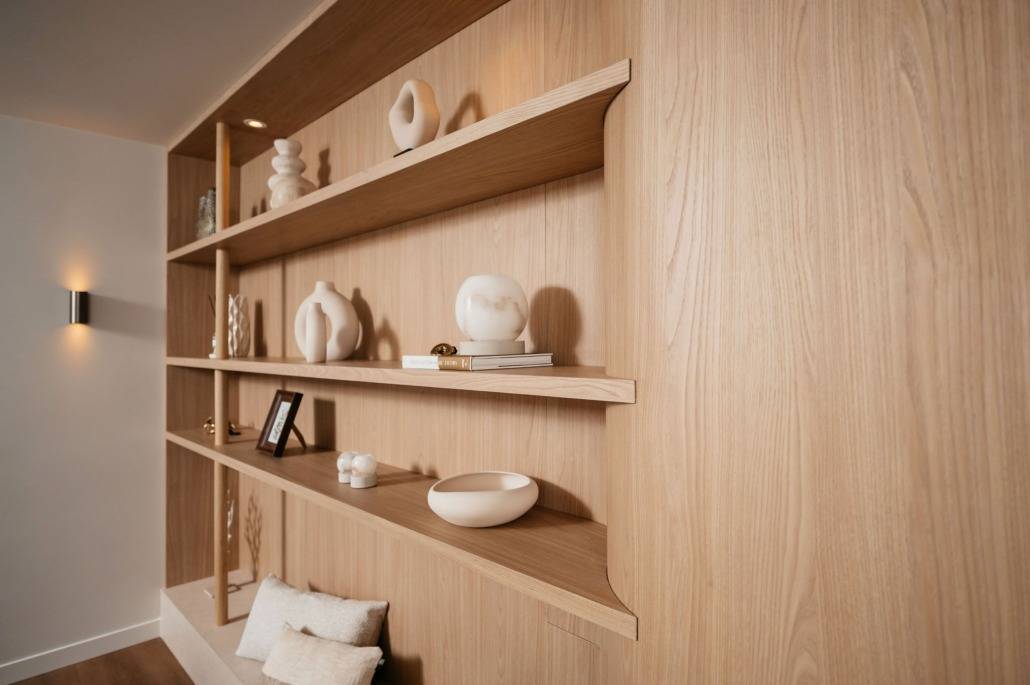 Minimalistische houten schappen met decoratieve witte keramiek, boeken, ingelijste foto's en kleine voorwerpen tegen een lichte houten wand. Kussens liggen opgestapeld op de vloer onder de planken.