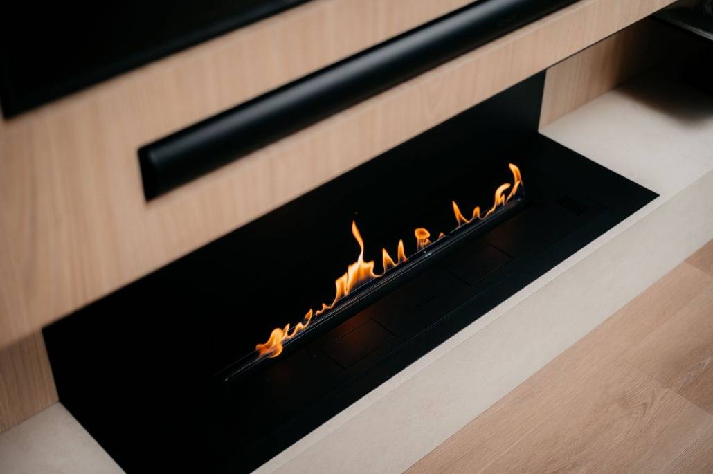 Een moderne lineaire haard met een smalle, horizontale vlam in een gestroomlijnd frame van zwart en licht hout, wat een minimalistische en gezellige uitstraling creëert.