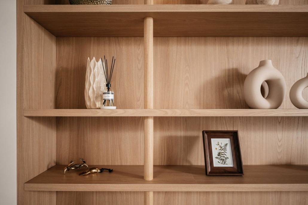 Een houten plank met decoratieve voorwerpen waaronder een gebeeldhouwde vaas, een rietverdamper, twee abstracte keramische vazen, een ingelijste botanische print en gouden glazen. De planken zijn spaarzaam gerangschikt in een minimalistische stijl.