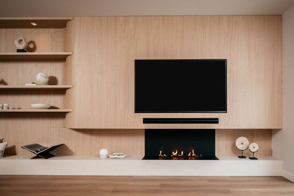 Een moderne woonkamer met een tv aan de muur boven een strakke zwarte open haard, licht houten lambrisering, ingebouwde planken met abstract decor en een lage witte plank met minimalistische ornamenten.