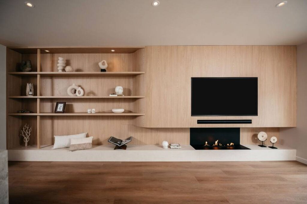 Moderne woonkamer met lichthouten ingebouwde planken met decoratieve voorwerpen, een flatscreentelevisie aan de muur en een strakke zwarte open haard eronder. De vloer is van hout en de ruimte ziet er minimalistisch en opgeruimd uit.