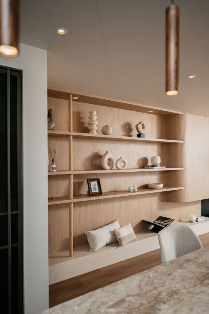 Minimalistische houten planken met neutraal gekleurde decoratieve vazen, schalen, abstracte sculpturen, een ingelijste foto en boeken. Kussens zijn geplaatst op een ingebouwde bank onder de planken, waardoor een gezellige, moderne hoek is ontstaan.