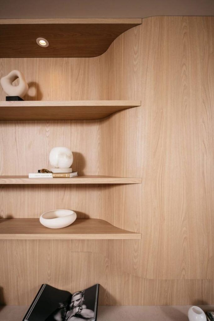 Minimalistische houten planken met abstracte witte sculpturen, een paar gestapelde boeken en een open zwart-wit fotoboek op een tafel. De achtergrond is licht houten lambrisering.