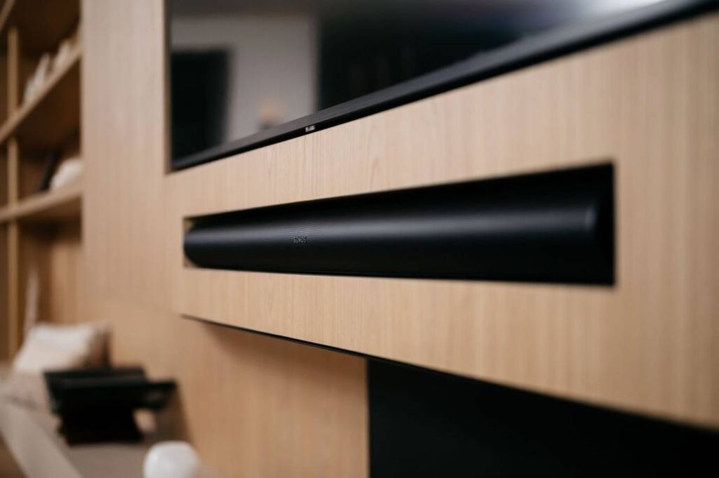 Close-up van een zwarte soundbar geïnstalleerd in een licht houten entertainmentmeubel onder een flatscreen-tv, met planken en decoratieve items op de achtergrond.