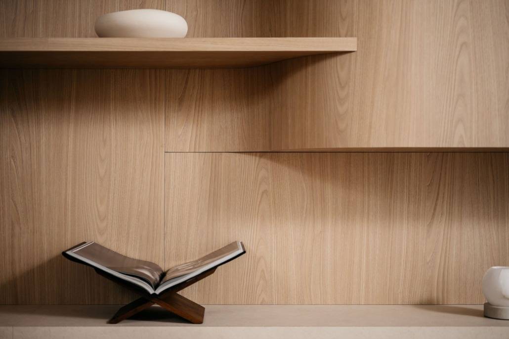 Een open boek op een houten standaard is geplaatst op een licht houten oppervlak, met een bijpassende houten muur en minimalistische planken met eenvoudig, modern decor op de achtergrond.