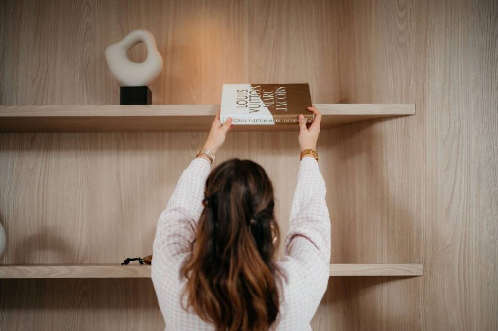 Een vrouw met lang bruin haar, van achteren gezien, plaatst een groot boek met de titel Louis Vuitton op een houten plank. Naast haar op de bovenste plank staat een wit abstract beeldhouwwerk.