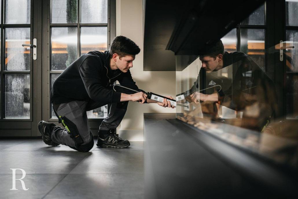 Een persoon knielt binnen bij een groot raam en gebruikt een kitpistool om de onderkant van een glazen wand of haard af te dichten. Reflecties zijn zichtbaar op het glasoppervlak. De scène is goed verlicht met natuurlijk licht.