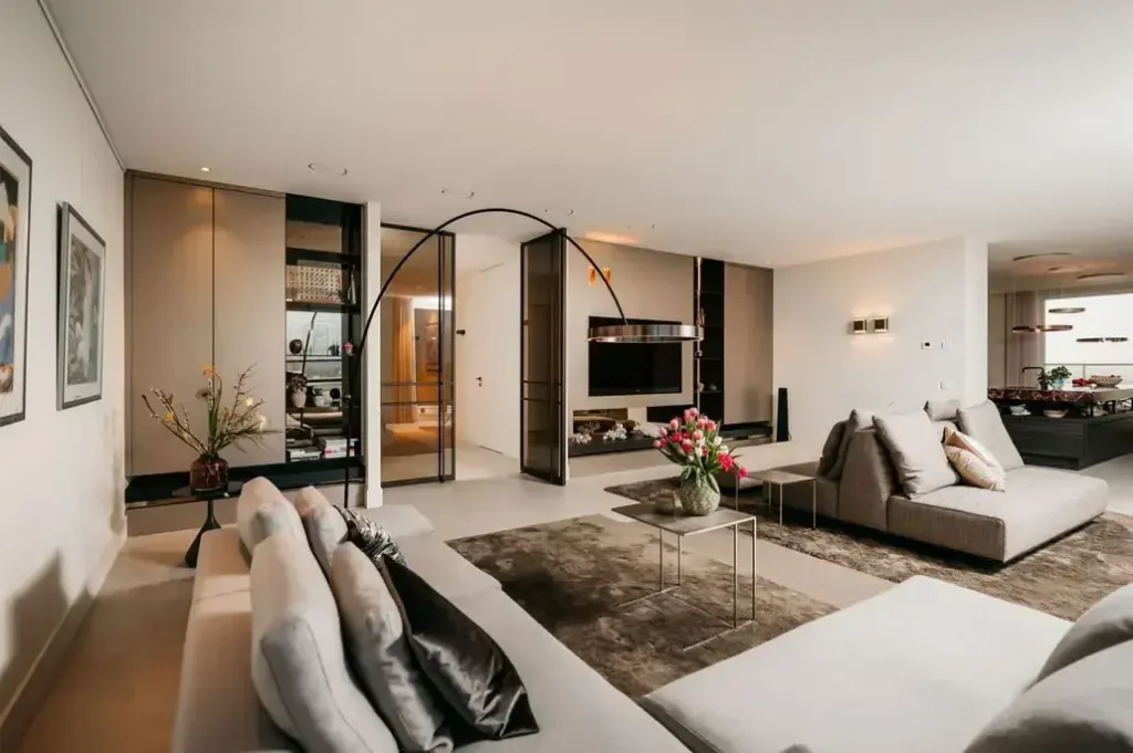 Moderne woonkamer met neutraal getinte banken, een groot vloerkleed, een salontafel met bloemen, muurkunst en een strakke tv. De ruimte is open, licht en stijlvol ingericht met eigentijds meubilair.