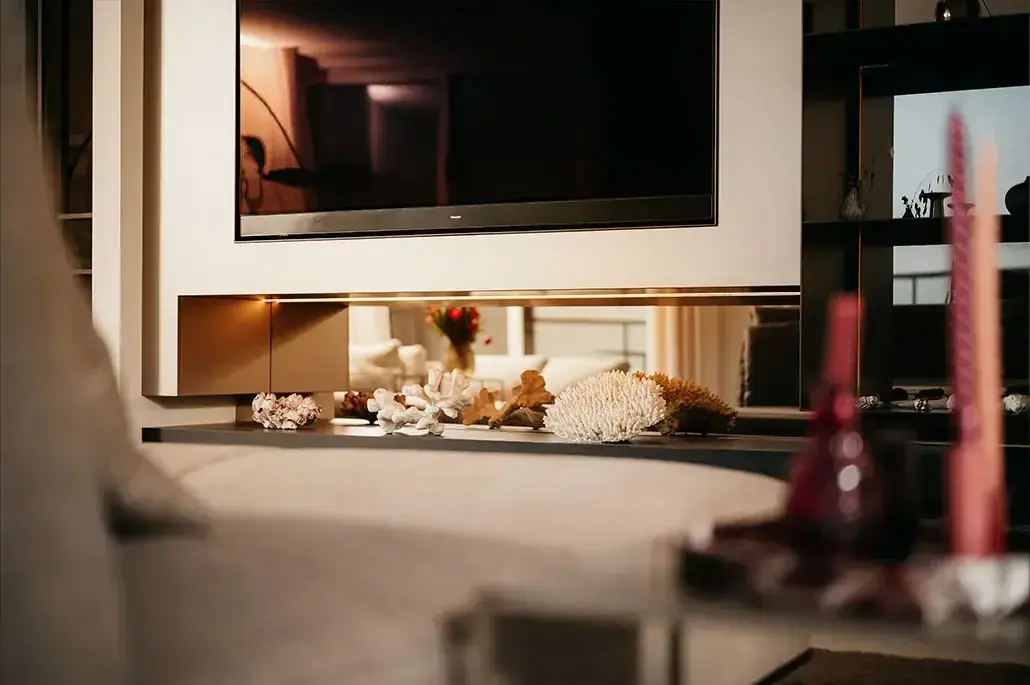 Een moderne woonkamer heeft een tv aan de muur boven een plank versierd met koraal en schelpen; op de voorgrond staat een vage salontafel met kaarsen.
