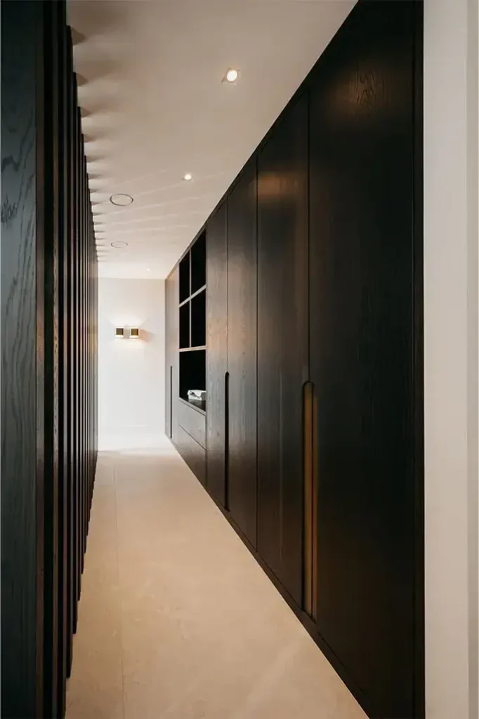 Een moderne hal met lichtbeige vloeren, donkerhouten lambrisering en kasten aan de ene kant, verticale houten latten aan de andere kant en ingebouwde plafondverlichting die een minimalistische, elegante look creëert.