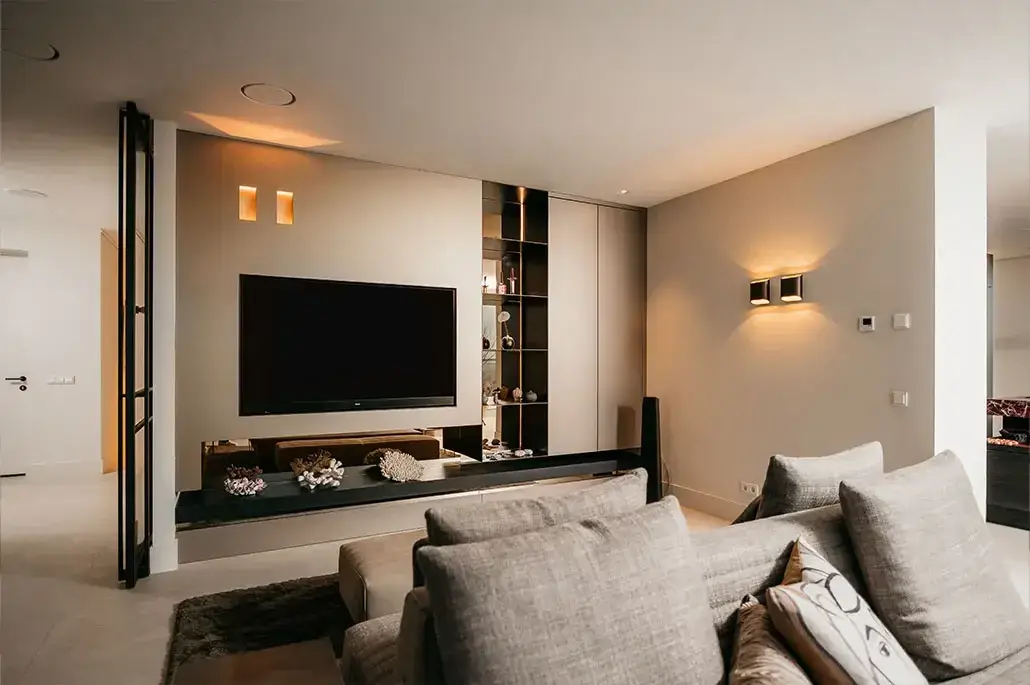 Moderne woonkamer met beige muren, een grote flatscreentelevisie aan de muur met ingebouwde planken, grijze sofa's met kussens en sfeervolle muurverlichting die een gezellige sfeer creëert.
