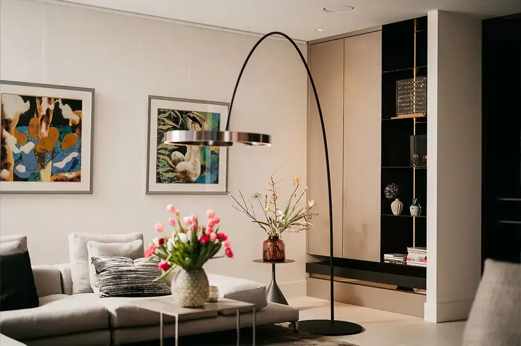 Een moderne woonkamer met een lichtgrijze sofa, kleurrijke kussens, een ronde tafel met een vaas roze tulpen, abstracte kunst aan de muur, een hoge booglamp en ingebouwde rekken met decoratieve items.