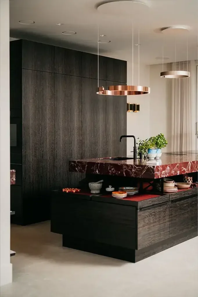 Moderne keuken met donkerhouten kasten, een groot eiland met een rood marmeren aanrechtblad, een zwarte kraan, koperen hanglampen en verschillende schaaltjes en kruiden op het aanrecht, waardoor de ruimte een stijlvolle, eigentijdse uitstraling heeft.