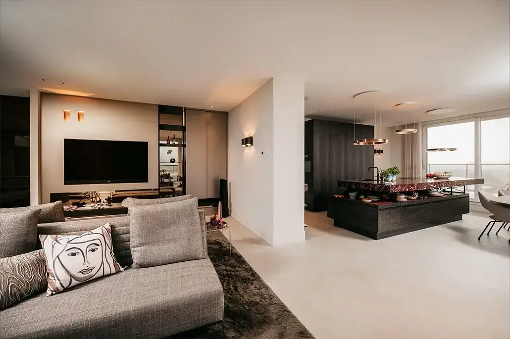 Moderne open woonkamer en keuken met neutrale tinten, een grijze sofa met een sierkussen met een gezichtsillustratie, tv aan de muur, minimalistische inrichting en grote ramen die natuurlijk licht binnenlaten.