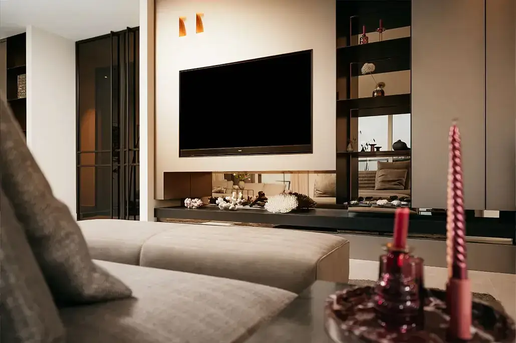 Een moderne woonkamer met een aan de muur gemonteerde flatscreentelevisie, ingebouwde planken met decoratieve voorwerpen, een lichtgekleurde bank en een salontafel met twee roze kaarsen en een glazen container.