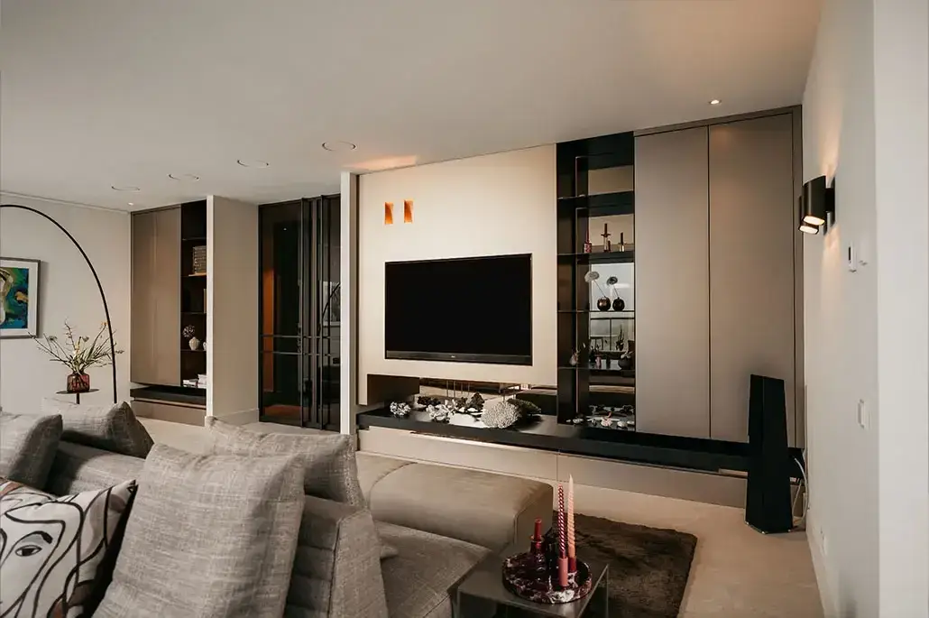 Een moderne woonkamer met een grote tv aan de muur, ingebouwde rekken met decoraties, een grijze sofa met patroonkussens en een kleine salontafel op een bruin vloerkleed. De ruimte heeft een minimalistische, neutraal getinte inrichting.