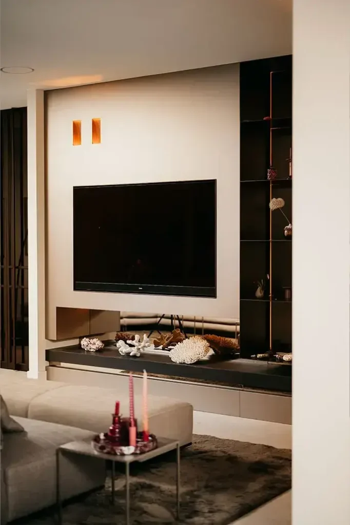 Een moderne woonkamer met een aan de muur gemonteerde flatscreentelevisie, ingebouwde rekken met decoratieve voorwerpen, een beige bank en een dienblad met roze kaarsen en glaswerk op een tafel. Warme verlichting creëert een gezellige sfeer.