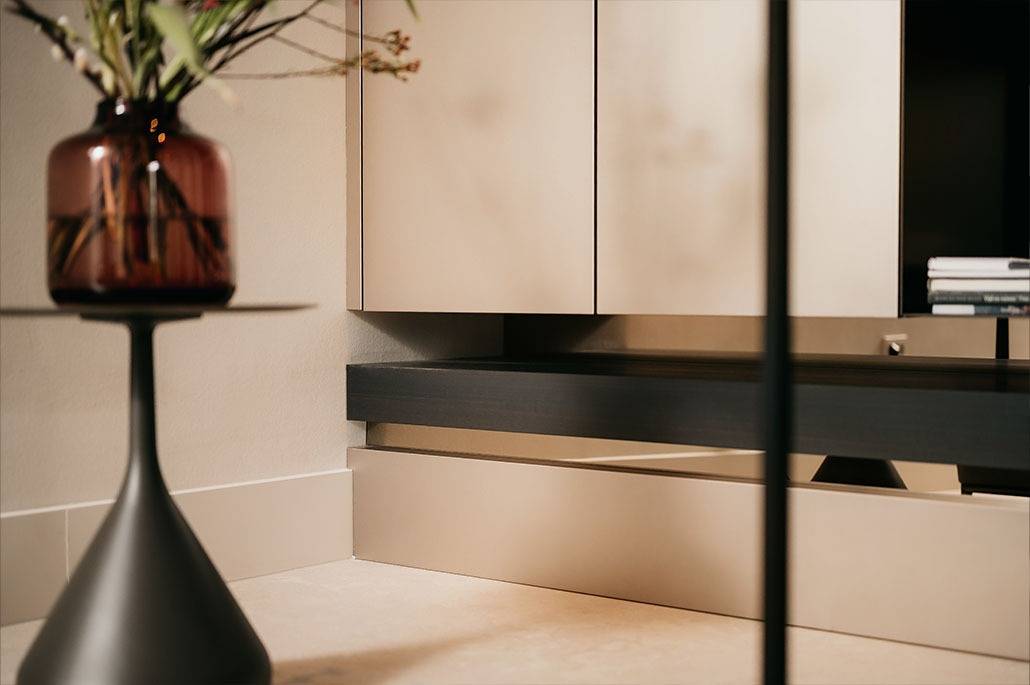 Een close-up van een moderne, minimalistische woonruimte met een ronde bijzettafel met een bruine glazen vaas en bloemen, lichtgekleurde kasten en een paar gestapelde boeken op een zwarte plank.