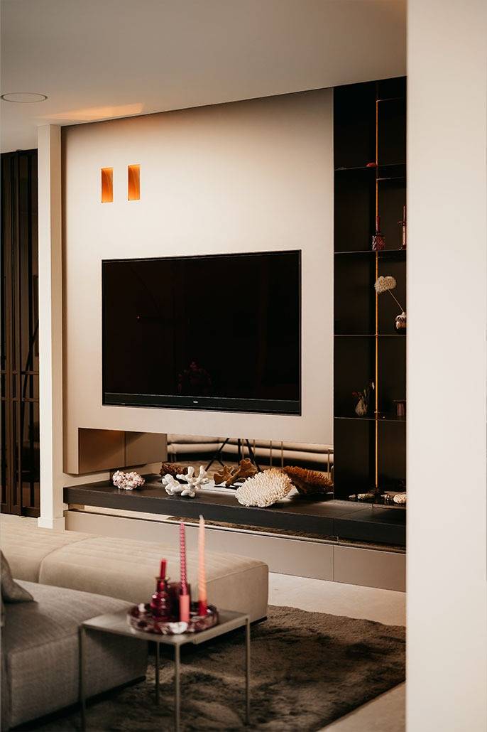 Moderne woonkamer met een tv aan de muur, ingebouwde rekken met decoratieve voorwerpen, zachte verlichting, een grijze bank en een salontafel met roze kaarsen op de voorgrond.