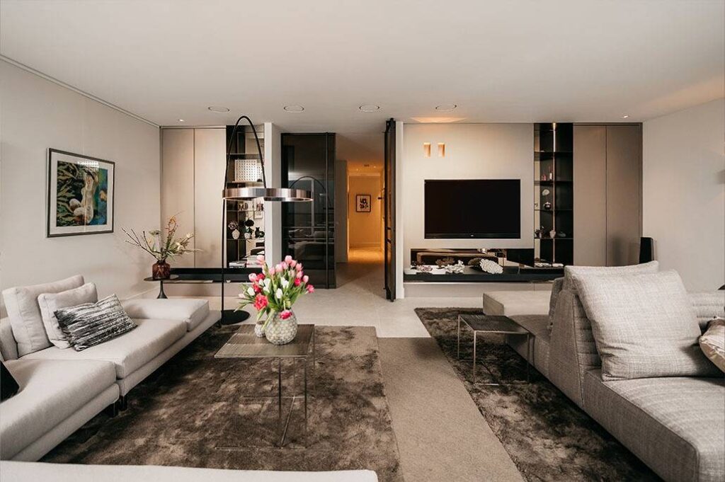 Moderne woonkamer met neutrale tinten, twee grijze banken, een glazen salontafel met roze tulpen, een grote tv aan de muur, donkere planken en een eigentijdse inrichting. Zachte verlichting creëert een gezellige, elegante sfeer.