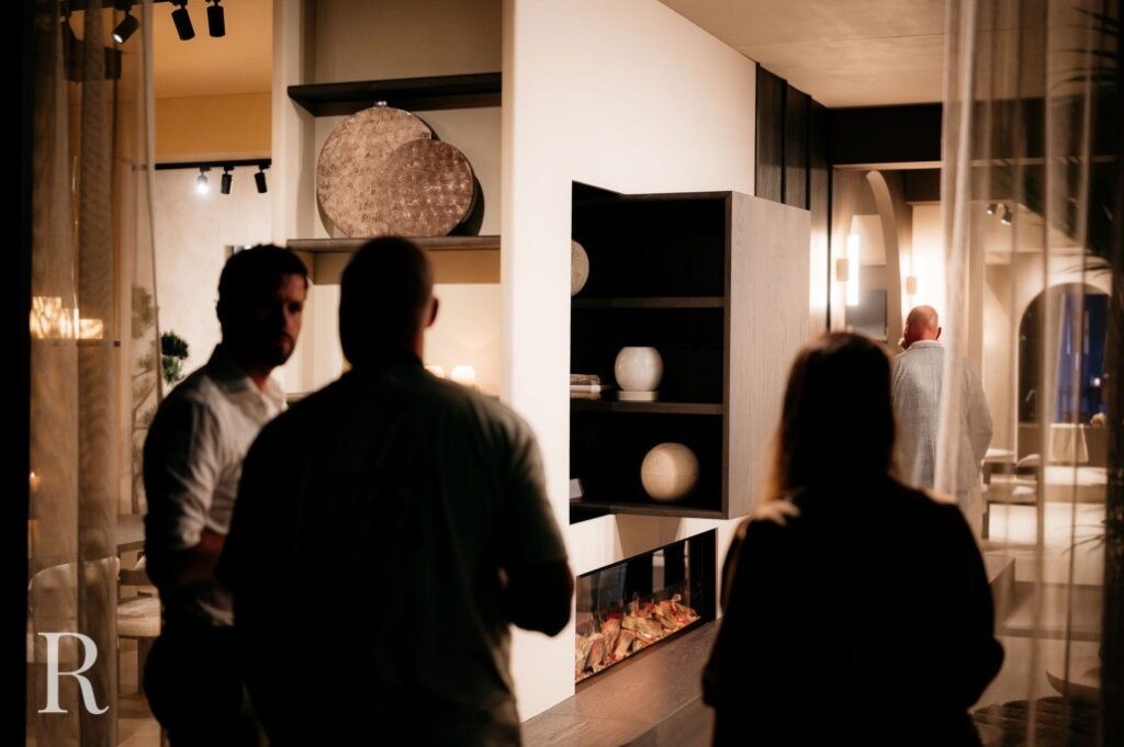 Drie mensen staan op de schemerige voorgrond van een moderne leefruimte, terwijl een vierde persoon wegloopt op de achtergrond. De kamer is voorzien van planken met decoratieve vazen en warme sfeerverlichting.