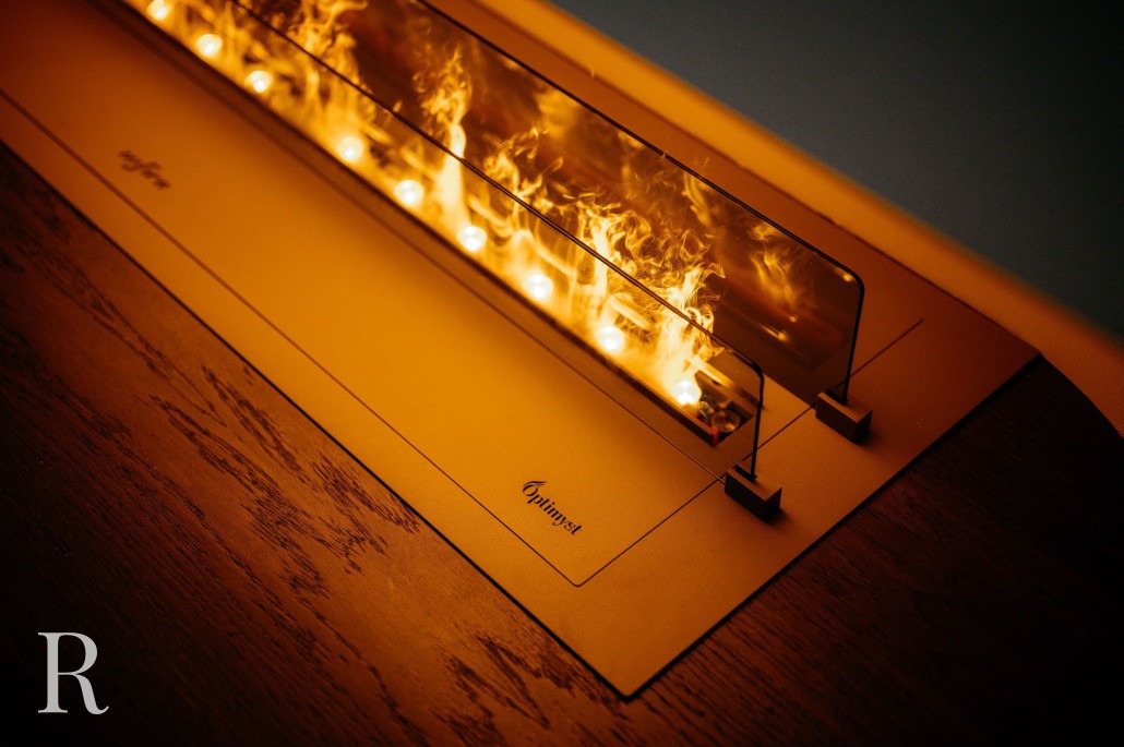 Een close-up van een moderne tafelhaard met een glazen barrière. Binnenin zijn feloranje vlammen zichtbaar die weerkaatsen op het glas en het houten oppervlak eronder verlichten. Het logo Element is afgedrukt op de voet.