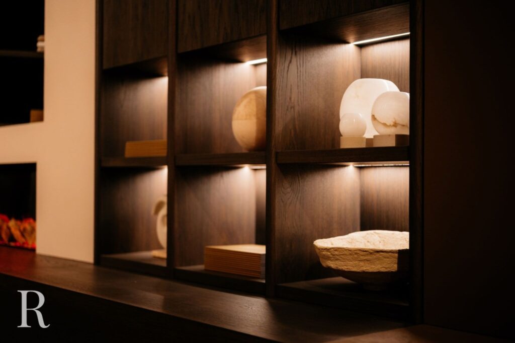 Een set donkere houten planken met ingebouwde verlichting toont minimalistische witte sculpturen en een schaal met ruwe textuur en creëert zo een warme, moderne sfeer.