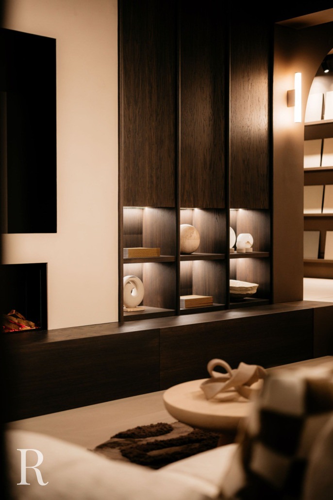 Een moderne woonkamer met donkerhouten planken met decoratieve voorwerpen, zachte verlichting en neutraal gekleurd meubilair. De ruimte heeft een warme, elegante sfeer met een ronde salontafel en abstracte decorstukken.