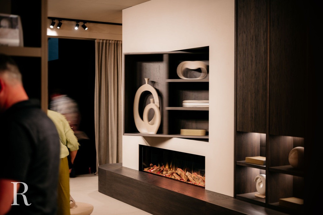 Een modern interieur met een ingebouwde elektrische open haard onder rekken met abstracte sculpturen. Twee mensen zijn gedeeltelijk zichtbaar aan de linkerkant en zachte verlichting creëert een gezellige sfeer. De letter R staat in de benedenhoek.