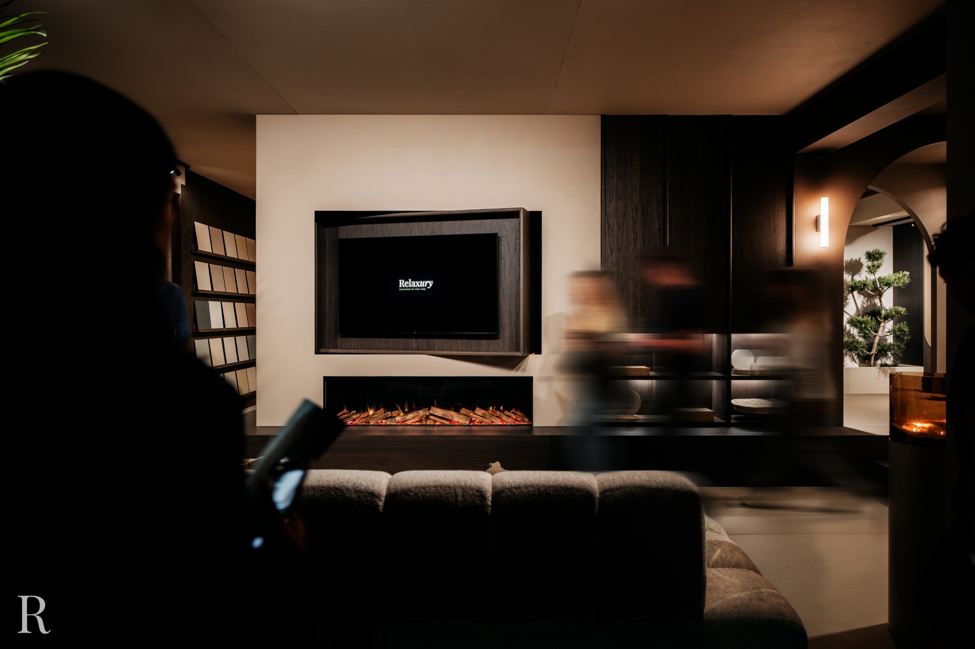 Een moderne woonkamer met een beige bank, een tv aan de muur boven een elektrische open haard, donkerhouten accenten en vage figuren die voorbij lopen; een persoon staat op de voorgrond en houdt een tablet vast.