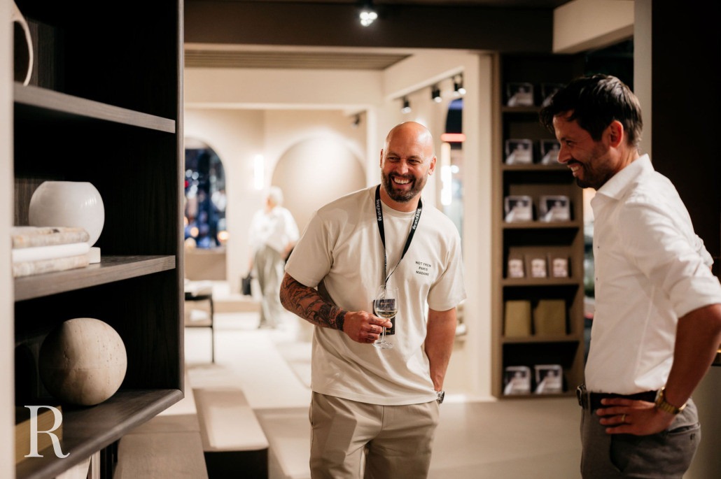 Twee mannen staan te praten en te glimlachen in een moderne, goed verlichte showroom of winkel. De ene man draagt een wit t-shirt en houdt een drankje vast, terwijl de andere een wit overhemd draagt. Aan de linkerkant zijn planken met decoratieve voorwerpen zichtbaar.