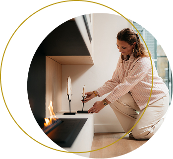 Een vrouw knielt bij een moderne open haard en past een decoratief veren ornament aan op een lage plank in een stijlvolle, hedendaagse woonruimte.