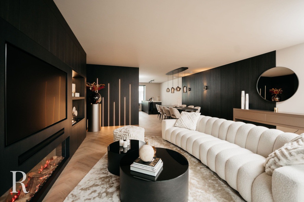 Moderne woonkamer met een gebogen witte bank, ronde zwarte salontafels, tv aan de muur, lineaire open haard en decoratieve vazen. De ruimte heeft donkere accentmuren, een grote ronde spiegel en een eethoek op de achtergrond.