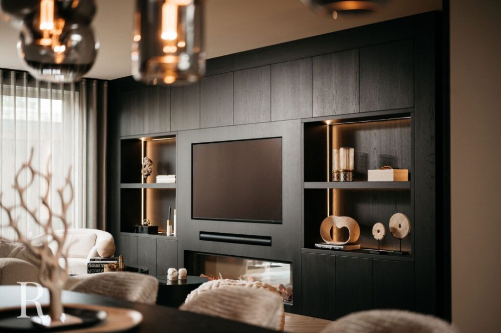Een moderne woonkamer met een donkerhouten inbouwwand met een grote flatscreentelevisie, planken met decoratieve voorwerpen en een lineaire elektrische open haard eronder. Zachte verlichting en neutraal gekleurd meubilair maken de gezellige ruimte compleet.