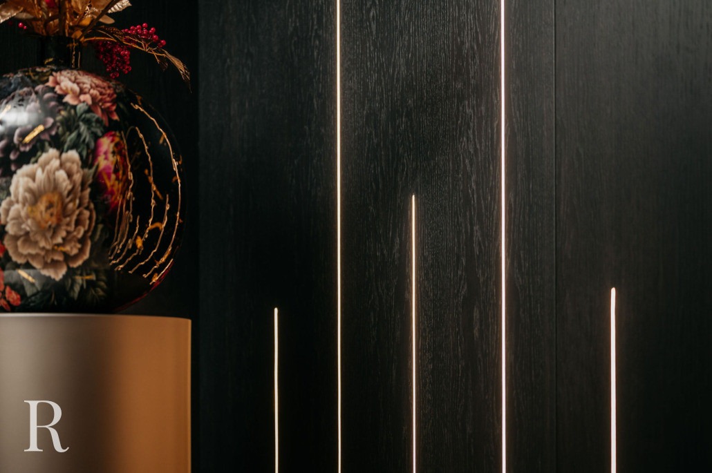Een decoratieve vaas met bloemmotieven staat op een beige sokkel naast een donkere houten muur met dunne, verticale verlichte lichtstroken. De letter R staat op de voorkant van de sokkel.