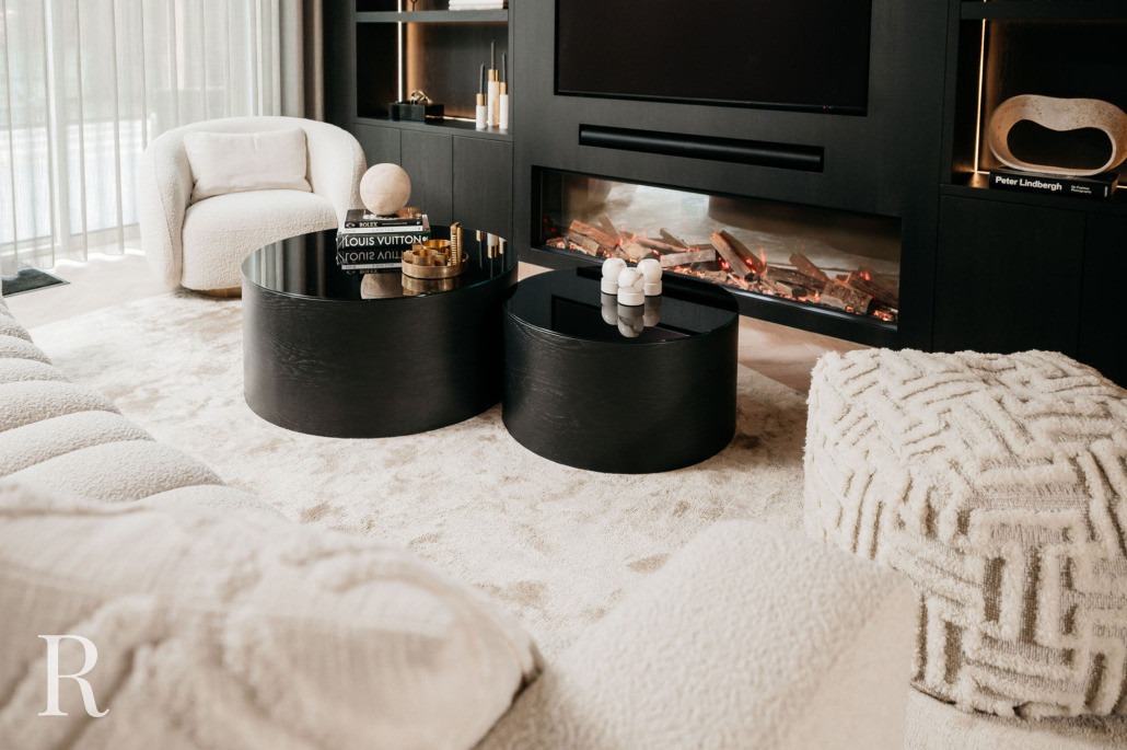 Een moderne woonkamer met gezellige crèmekleurige stoelen, twee ronde zwarte salontafels, een voetenbank met geometrische textuur, een open haard en ingebouwde planken met decoratieve voorwerpen, alles op een zacht, licht tapijt.