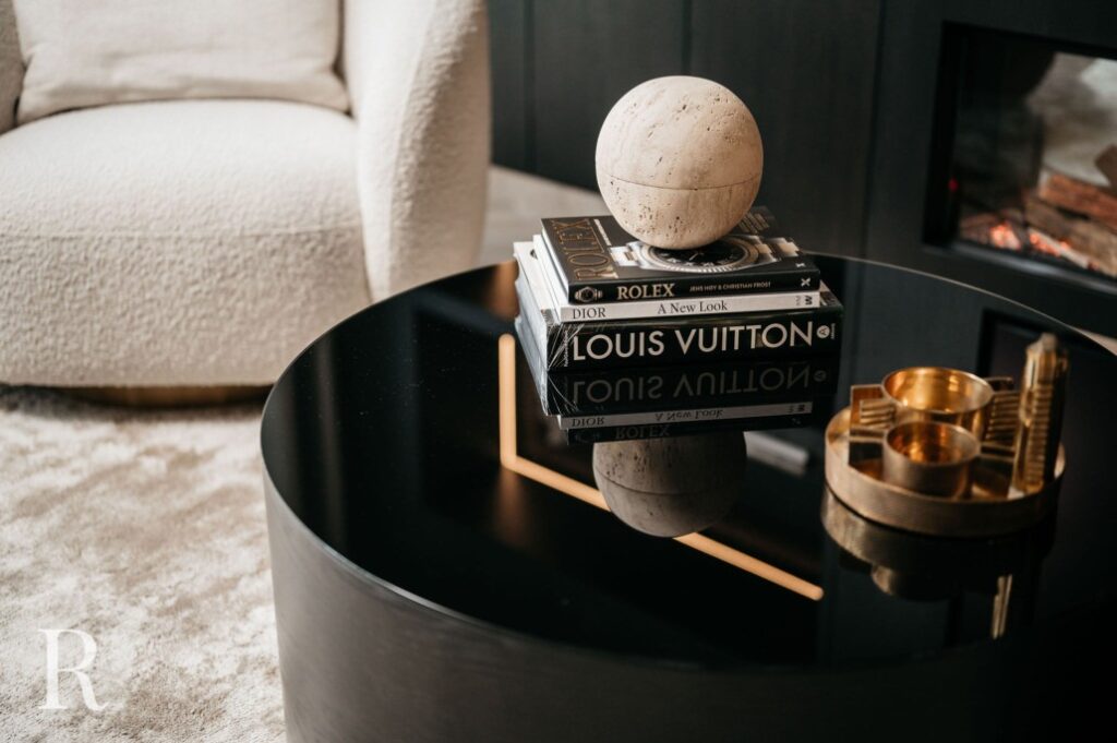 Een moderne zwarte salontafel met gestapelde modeboeken, een rond beige decoratief object erop, een gouden dienblad met kaarsenhouders, een crèmekleurige fauteuil en een open haard in een stijlvolle woonkamer.