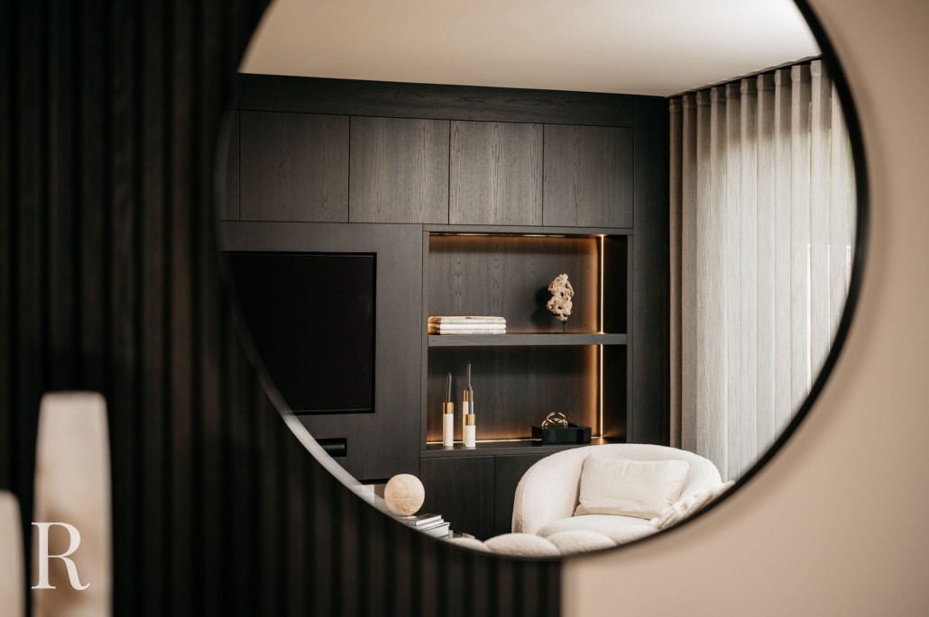 Spiegeling in een ronde spiegel die een moderne woonkamer laat zien met donkerhouten lambrisering, een ingebouwde plank met decor, een tv aan de muur en een beige fauteuil door kamerhoge gordijnen.