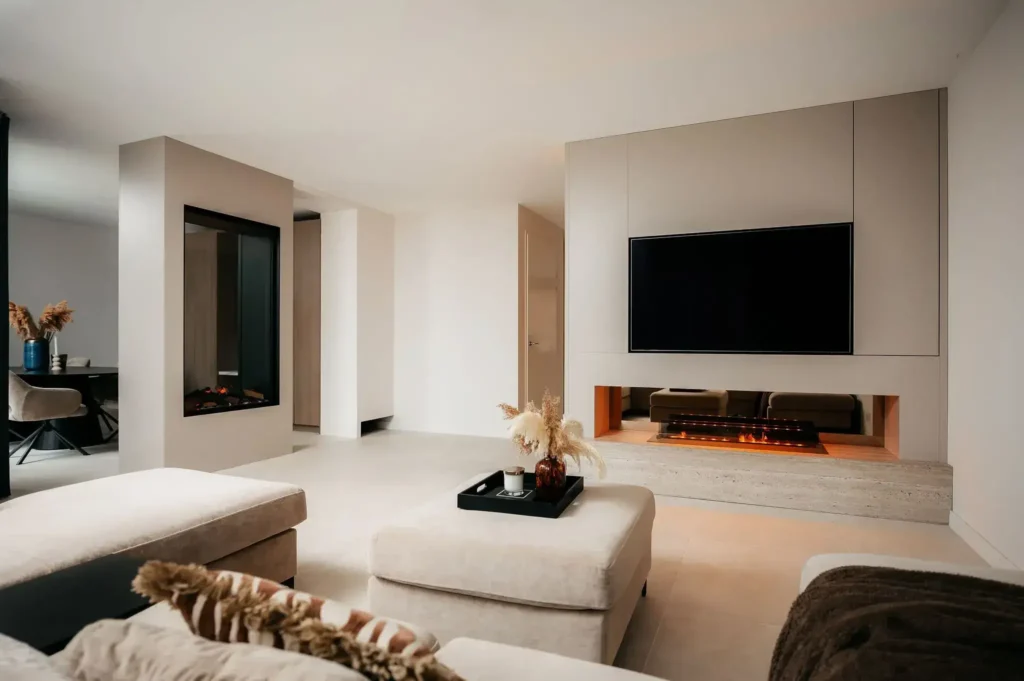 Moderne woonkamer met minimalistische inrichting, met een grote tv op een stijlvolle cinewall boven een ingebouwde elektrische open haard, neutraal gekleurd meubilair en een gezellige zithoek met een salontafel en een arrangement van gedroogd pampagras.