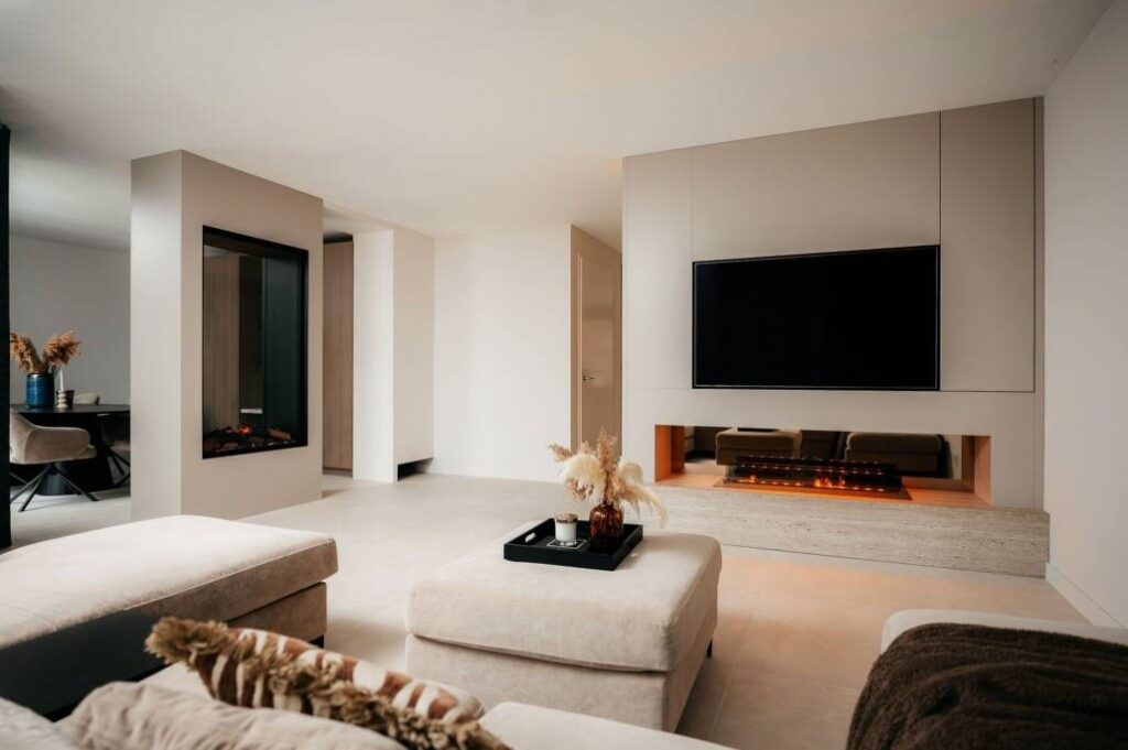 Moderne woonkamer met neutrale tinten, met een stijlvolle cinewall met een tv aan de muur boven een ingebouwde elektrische open haard, een grote voetenbank, zachte stoelen en een minimalistische inrichting met gedroogde planten. Warme verlichting creëert een gezellige sfeer.