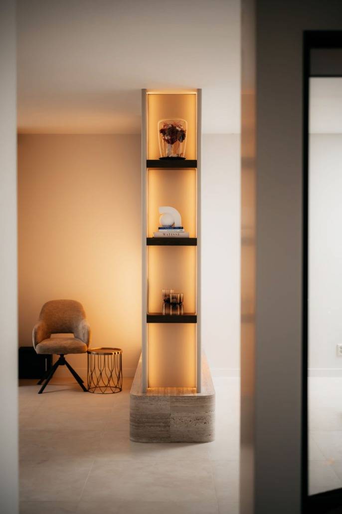 Een smalle, ingebouwde stelling met warme verlichting lijkt op een cinemamuur en toont decoratieve voorwerpen zoals een vaas en abstracte sculpturen. Op de achtergrond maken een gezellige fauteuil en een ronde bijzettafel de moderne, minimalistische kamer compleet.