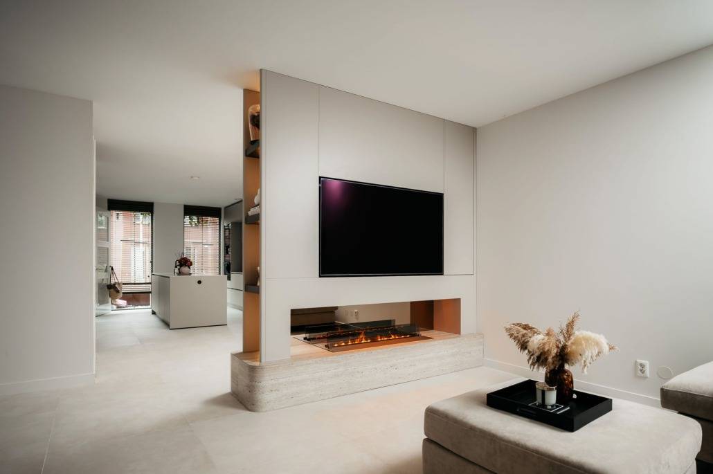 Een moderne woonkamer met een cinewall met een tv aan de muur boven een ingebouwde elektrische open haard, muren en vloeren in neutrale tinten, een minimalistische inrichting en een dienblad met gedroogde planten op een beige voetenbank.