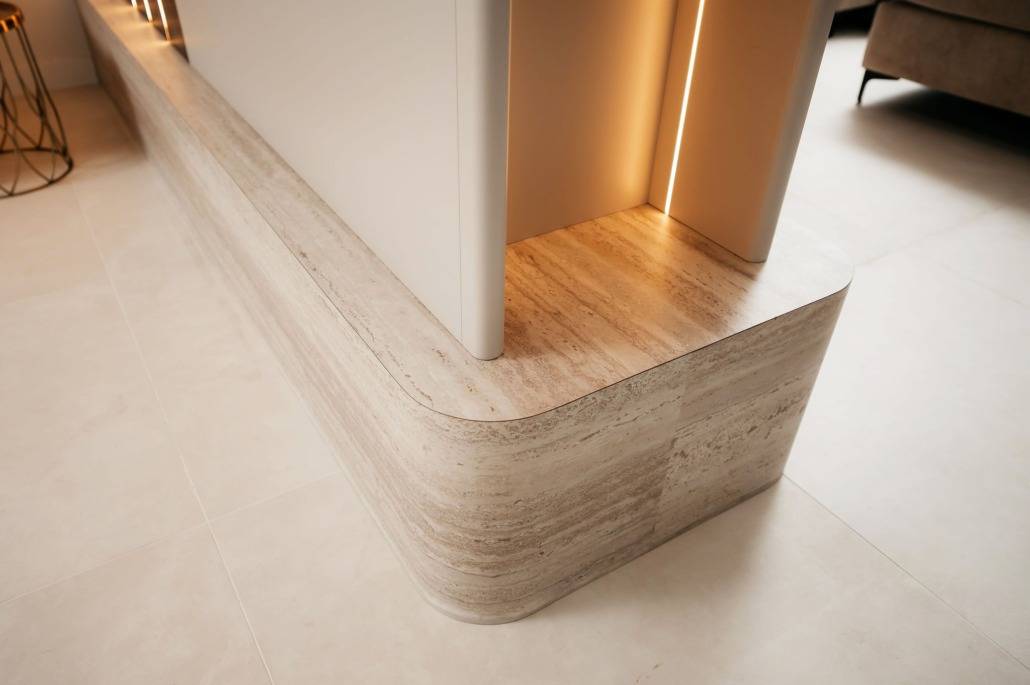 Een close-up van een modern interieur met een afgeronde hoek, licht houten of stenen basis en een wit paneel erboven dat lijkt op een stijlvolle cinewall, verlicht door warme verzonken verlichting.