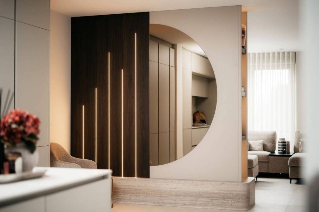 Een moderne leefruimte is voorzien van een grote ronde spiegel in een cinewall met verticale LED-lichtstrips, een beige fauteuil en neutraal gekleurd meubilair, met natuurlijk licht door de transparante gordijnen.