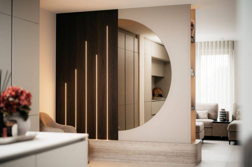 Een moderne leefruimte is voorzien van een grote ronde spiegel in een cinewall met verticale LED-lichtstrips, een beige fauteuil en neutraal gekleurd meubilair, met natuurlijk licht door de transparante gordijnen.